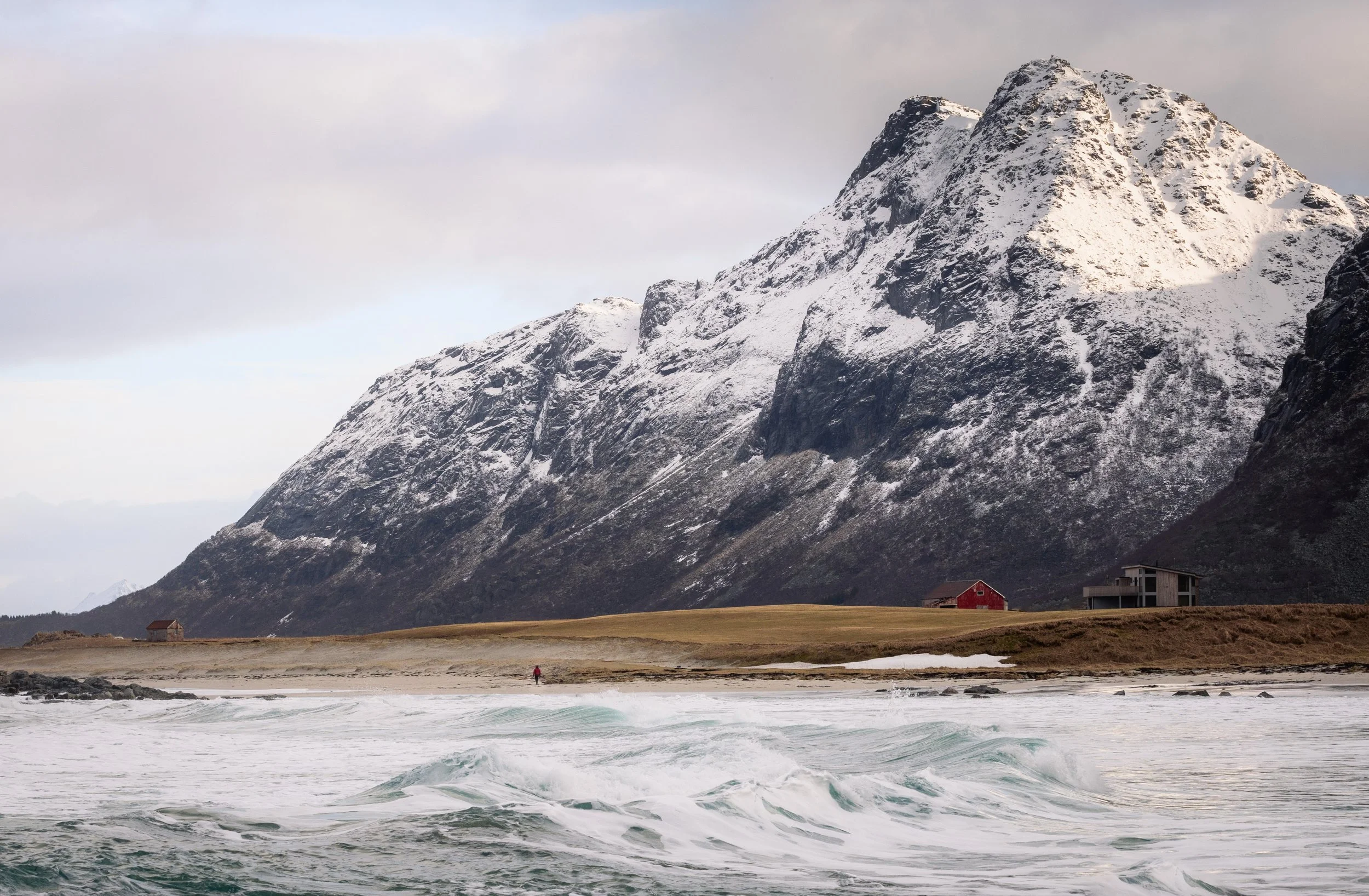 Surf running below a snow-covered massif with farm buildings along the shore, Lofoten, Norway — the peaks are ancient gneiss, among the oldest rock in Europe.