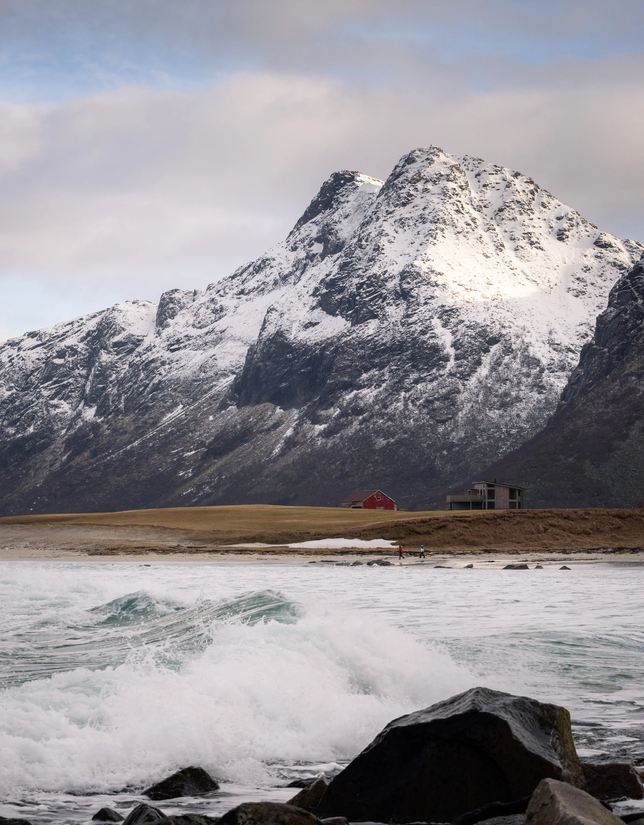 Surf breaking below a snow-covered peak with a red farmhouse on the shore, Lofoten, Norway — people have farmed and fished this coast for over a thousand years.