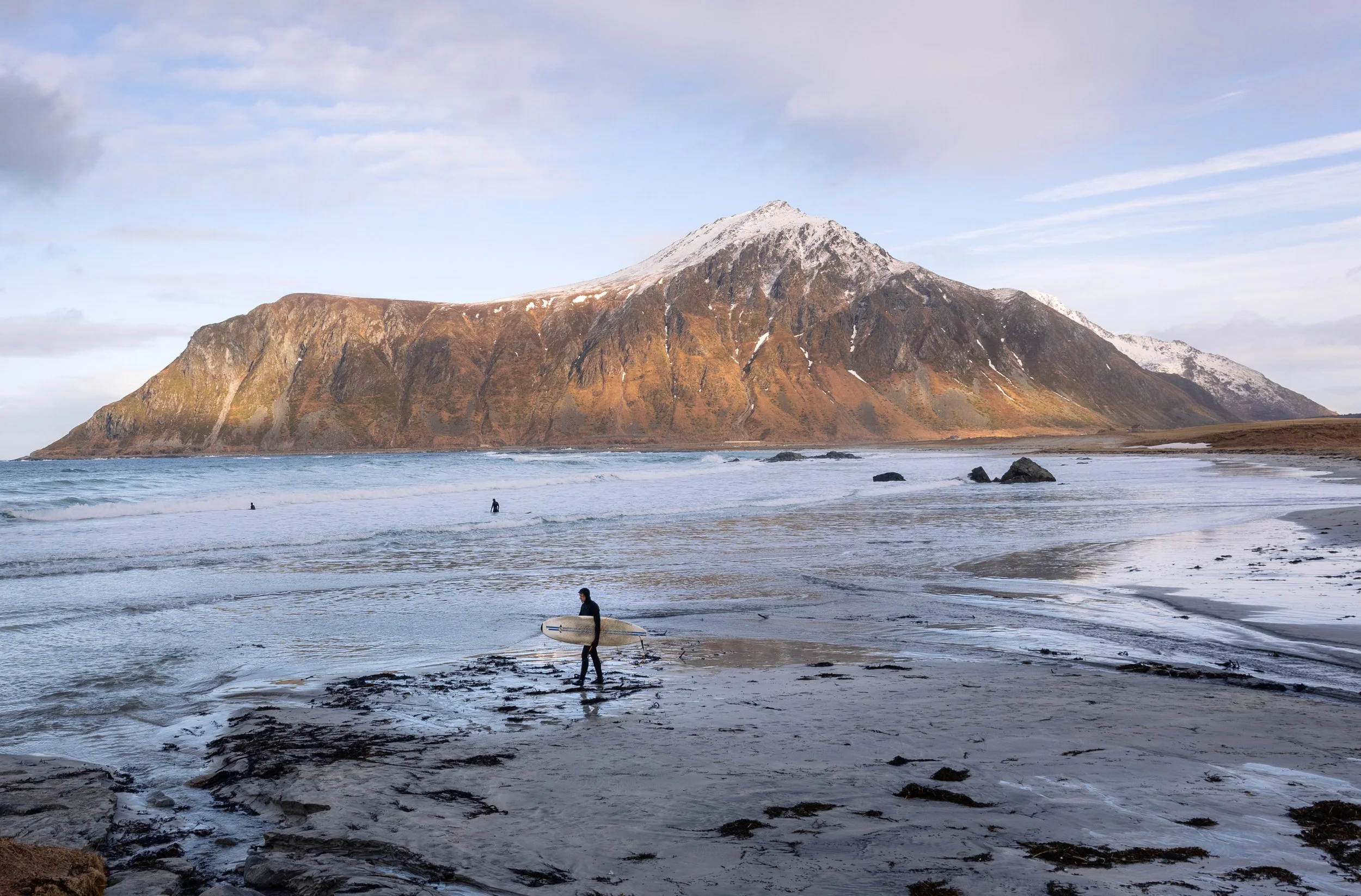 A surfer standing on the shoreline with the peak filling the bay behind, Lofoten, Norway