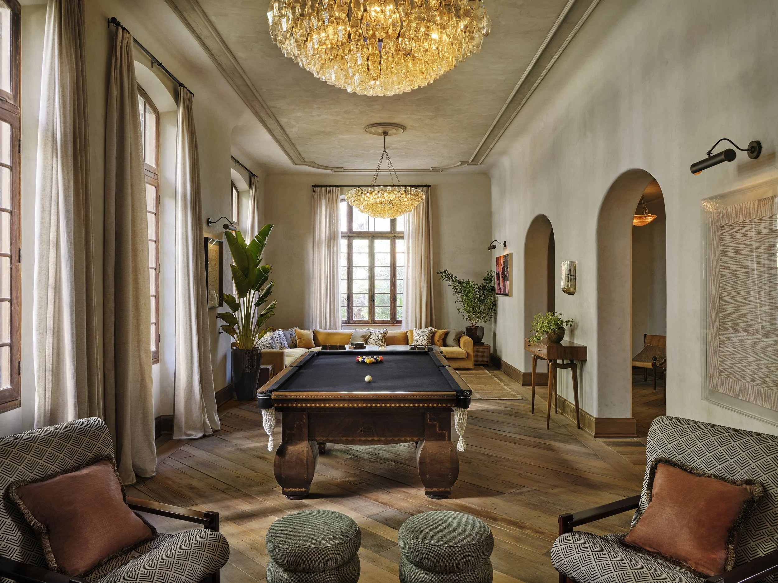 Living room with large windows, beige curtains, a chandelier, a pool table in the center, plants, artwork on the wall, and seating area with patterned chairs and cushions.
