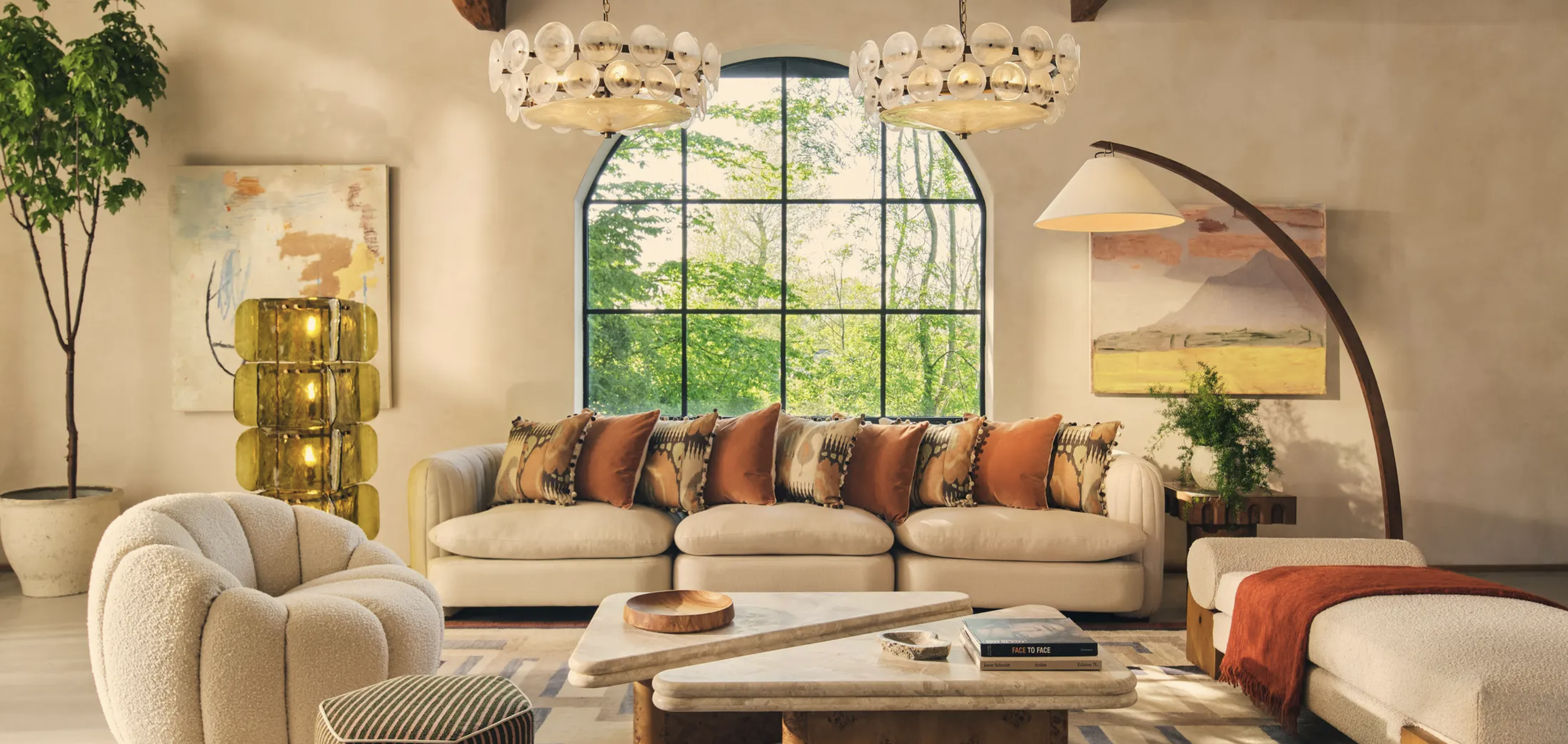 Living room with large arched window, beige sofa with patterned cushions, cream armchair, marble coffee table with books, potted plant, yellow stack lamp, abstract artwork, and greenery visible outside.