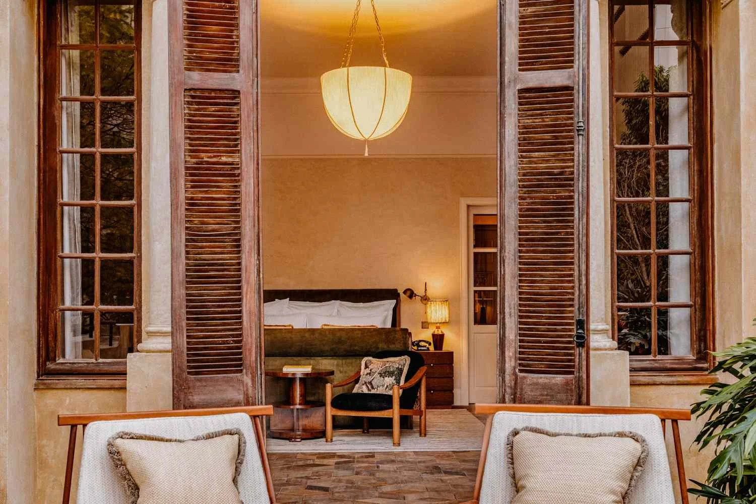 View of a cozy interior room with a bed, wooden side table, lamp, and a sitting chair, seen through large open wooden window shutters.