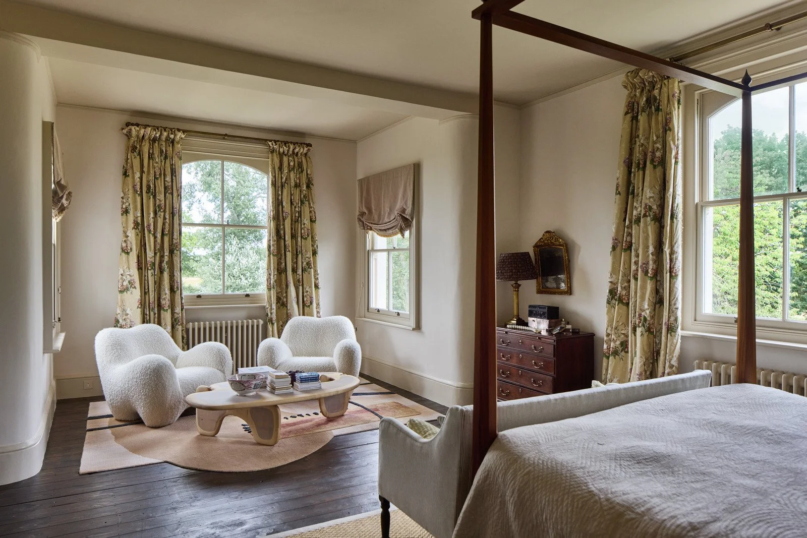 A vintage bedroom with a four-poster bed, cream upholstery, a wooden chest of drawers, a lamp, and partially open floral curtains revealing a large window with green trees outside.