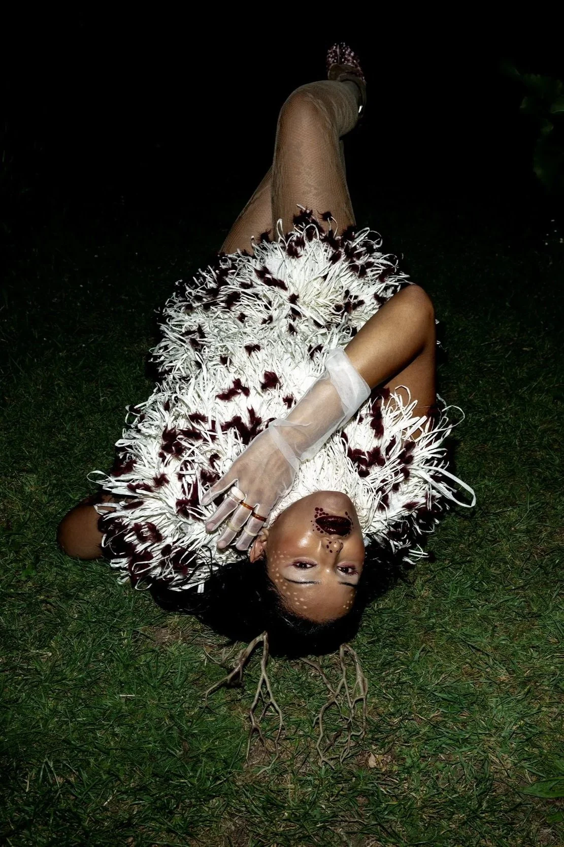 A woman lying on grass wearing a feathered white dress with brown spots, transparent gloves, and antler headband, with makeup resembling a deer and face paint resembling tree branches.