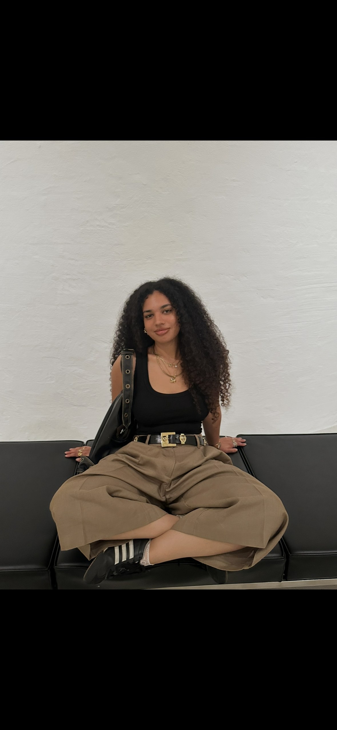 Young woman with curly hair sitting on a black bench against a white wall, wearing a black sleeveless top, khaki pants, black sneakers, and gold jewelry.
