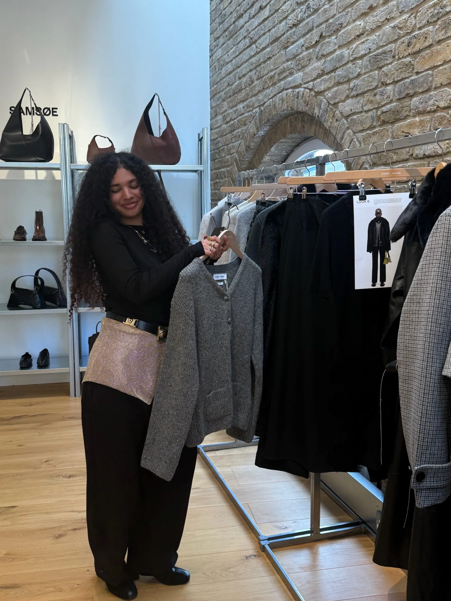 A woman with curly hair shopping for clothes in a boutique store, holding a gray sweater, with handbags and shoes displayed on shelves in the background.