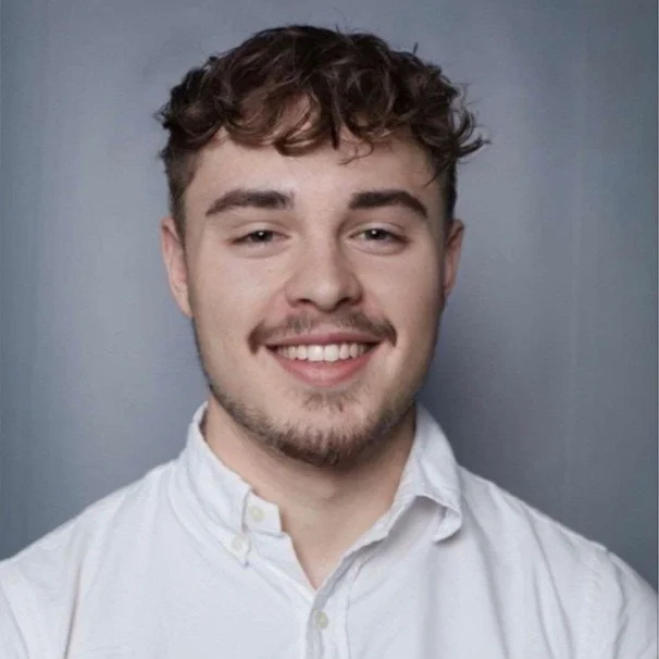 A young man with curly brown hair, light facial hair, smiling, wearing a white button-up shirt, standing against a gray background.