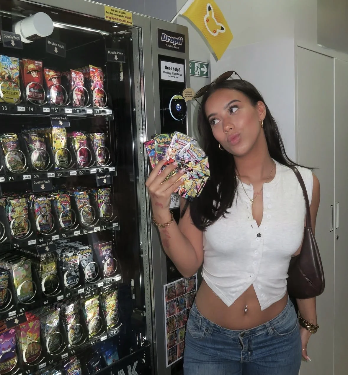 A young woman standing beside a vending machine, holding several Pokémon trading cards, with sunglasses on her head and wearing a white sleeveless button-up crop top and jeans.