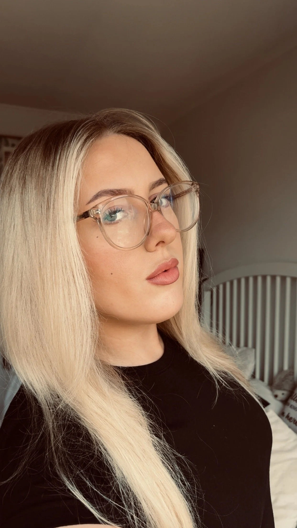 A woman with long blonde hair, wearing large clear glasses and a black top, looking to the side indoors.