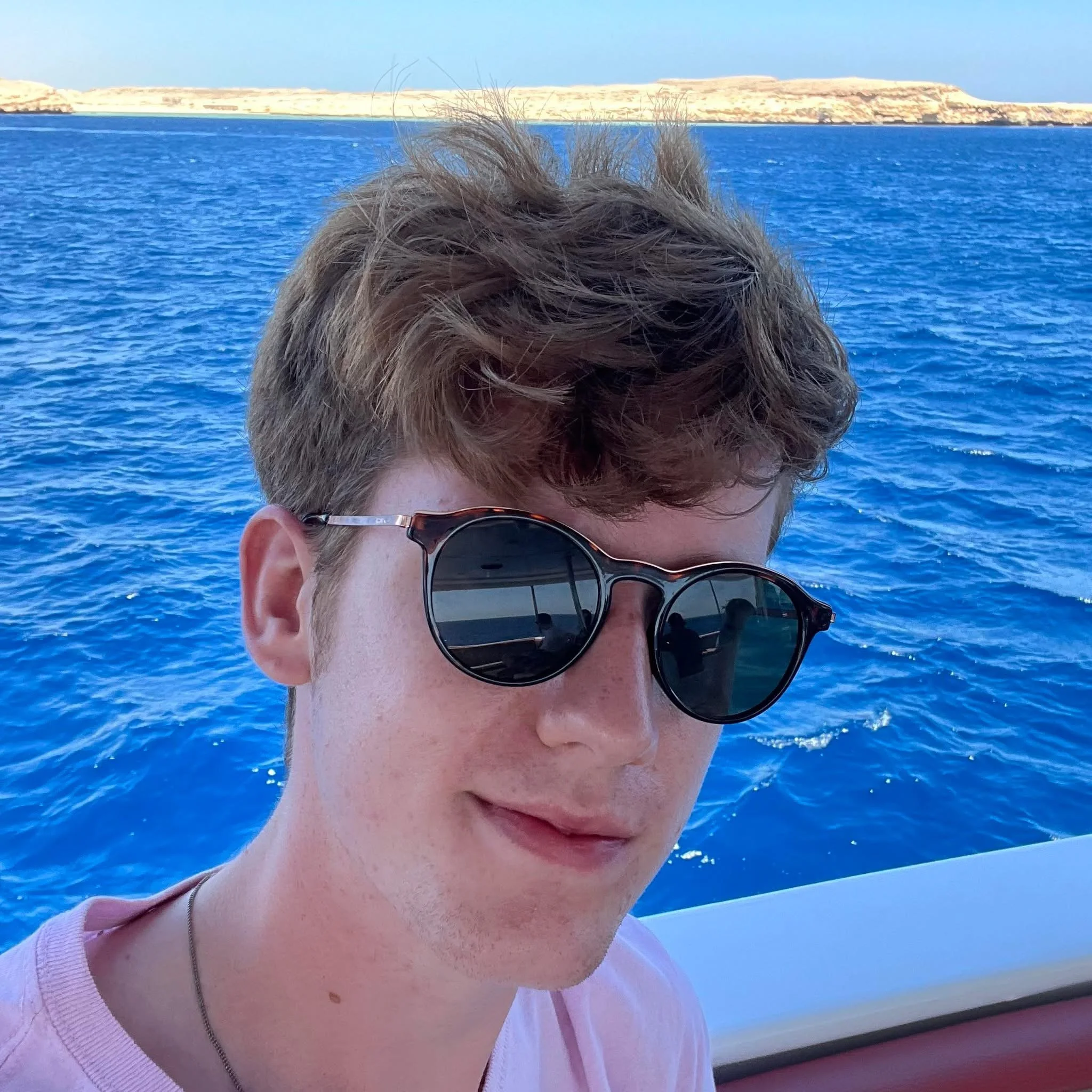 Close-up of a person with short curly hair wearing sunglasses on a boat, with blue ocean water and rocky landscape in the background.