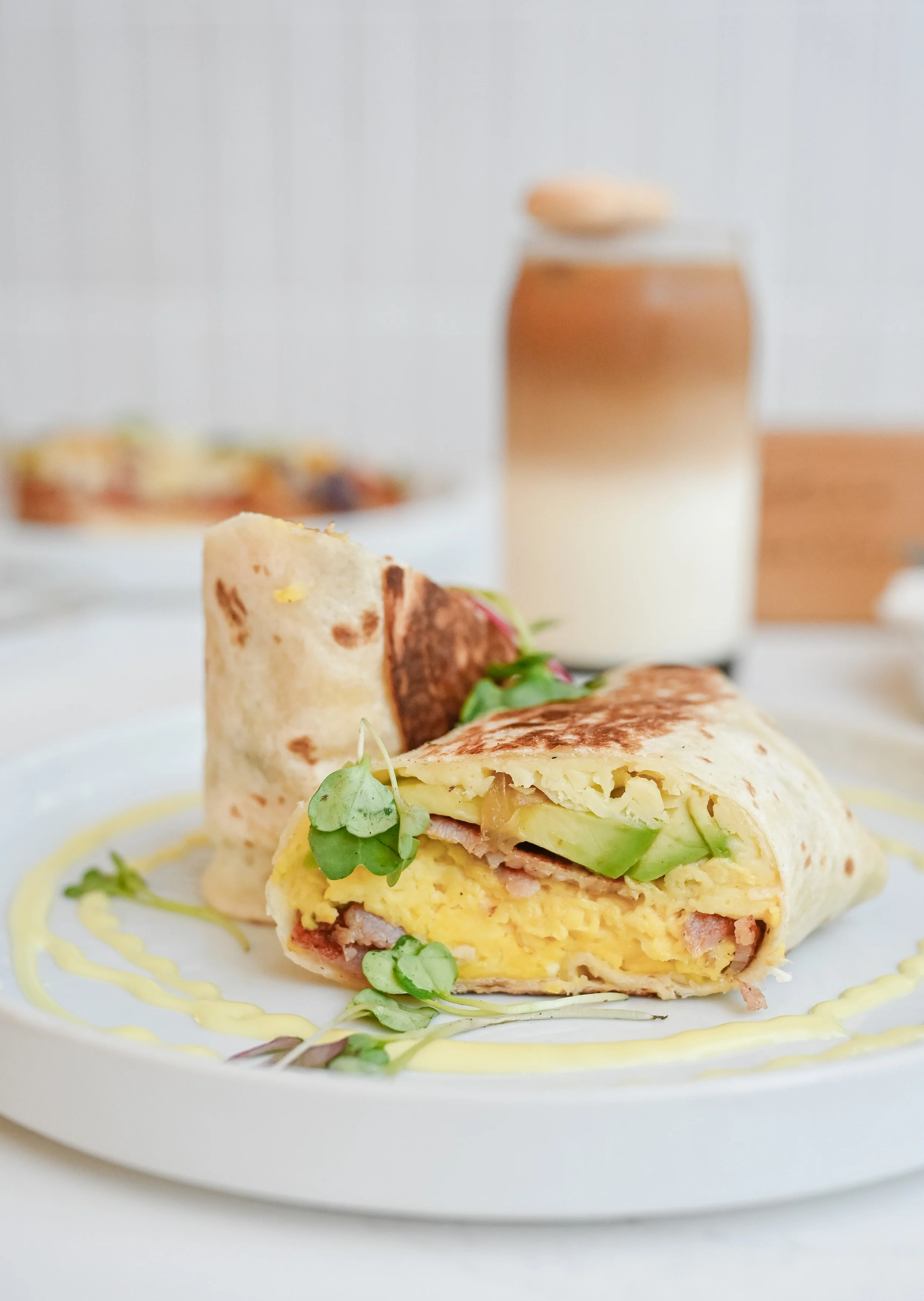 A close-up of a breakfast burrito cut in half on a white plate, garnished with microgreens and a drizzle of yellow sauce, with a blurred plate of food and a jar of chocolate milk in the background.