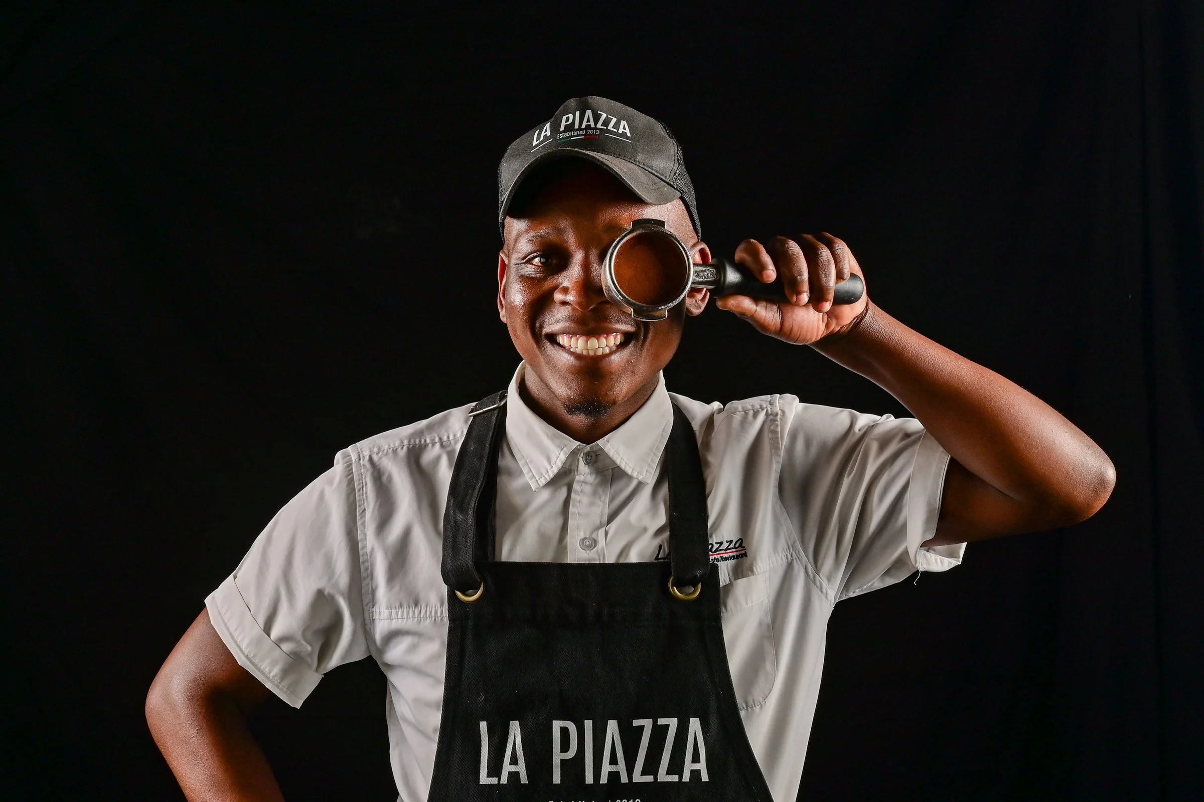 A smiling male chef holding a coffee mug to one eye, wearing a black cap with "LA PIAZZA" written on it, a white shirt, and a black apron with "LA PIAZZA" printed on it against a black background.