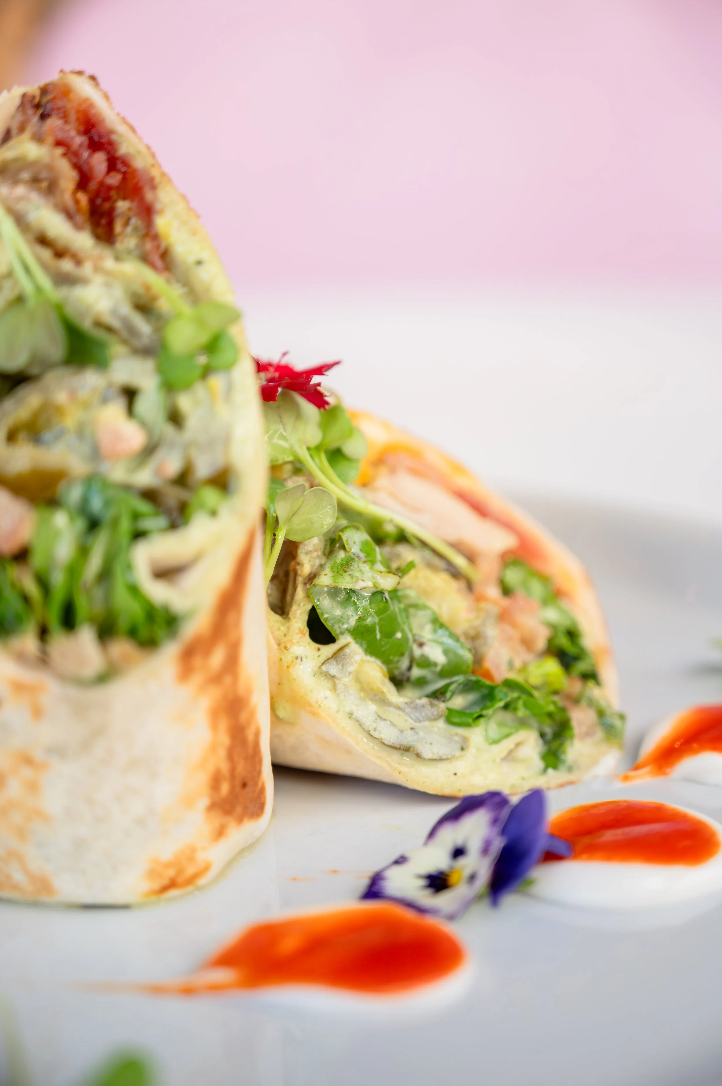 Close-up view of a wrap sandwich filled with greens, vegetables, and creamy sauce, garnished with edible flowers and drizzled with sauces on a white plate against a pastel background.