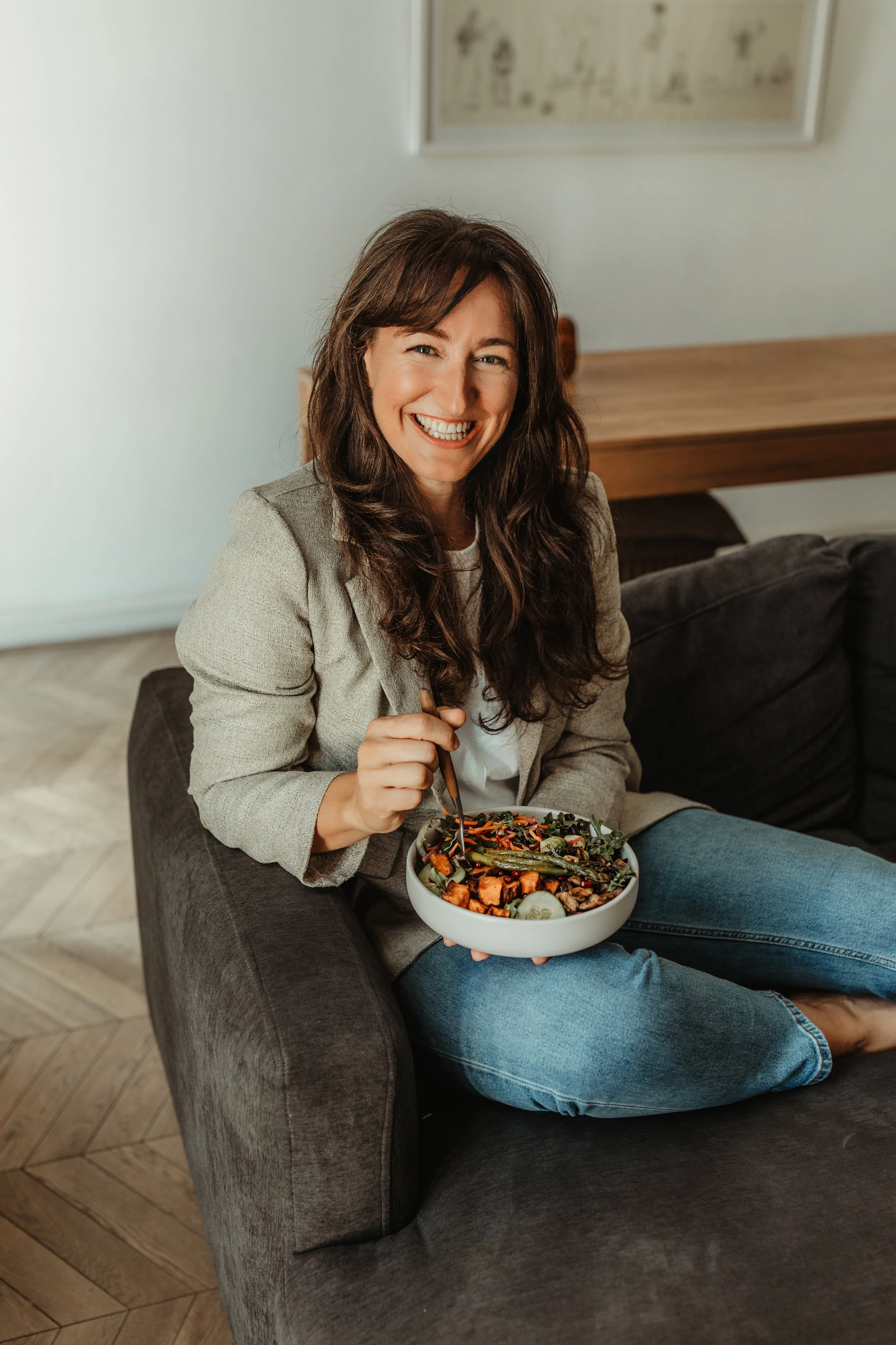A woman with brown hair and a beige blazer sitting on a gray couch, smiling while holding a bowl of salad. The background has a wooden table and framed art on a white wall.