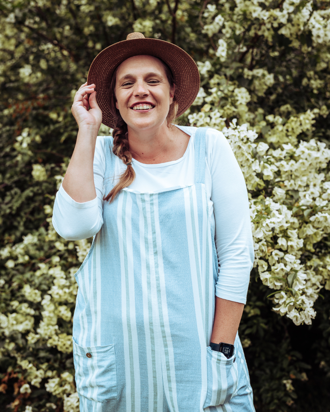 A smiling woman wearing a straw hat, a light blue and white striped dress over a white long-sleeve shirt, standing in front of white flowering bushes.