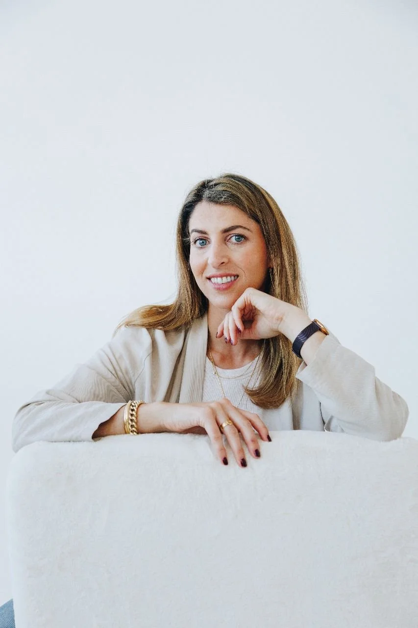 A woman with brown hair and blue eyes smiling while leaning on a white surface, dressed in a beige blazer and accessorized with jewelry, against a plain white background.