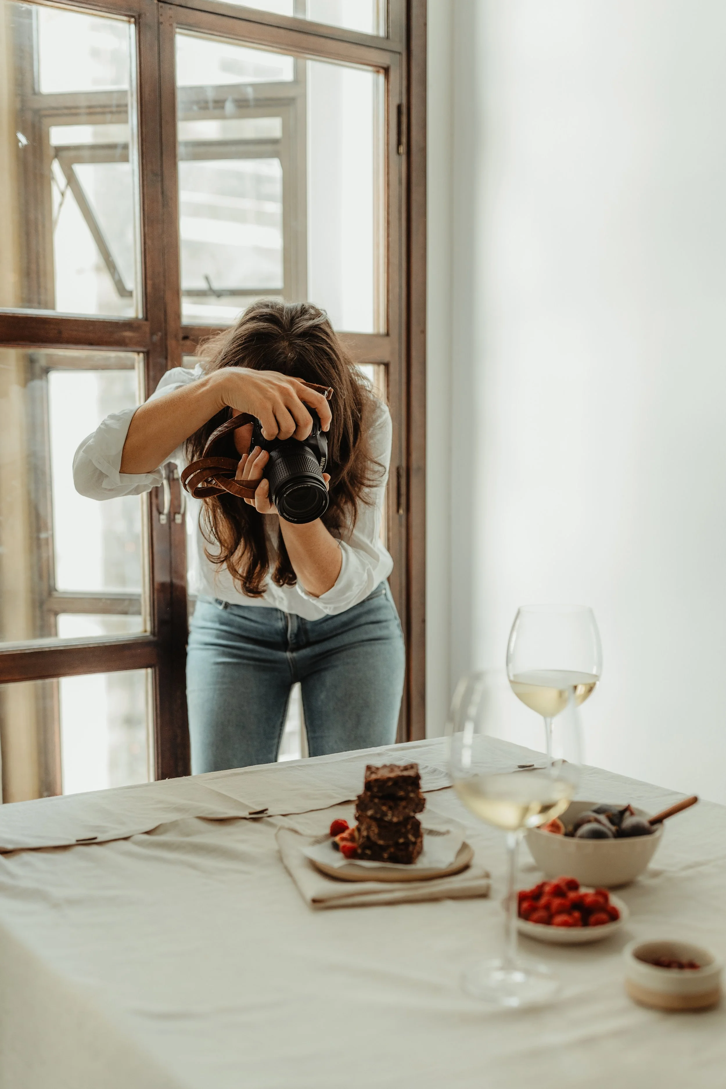 A woman with wavy brown hair wearing a white shirt and blue jeans taking a photo with a camera in a bright room with large wooden-framed windows. In the foreground, there is a table set with a white tablecloth, a stack of dessert bars, a bowl of stra