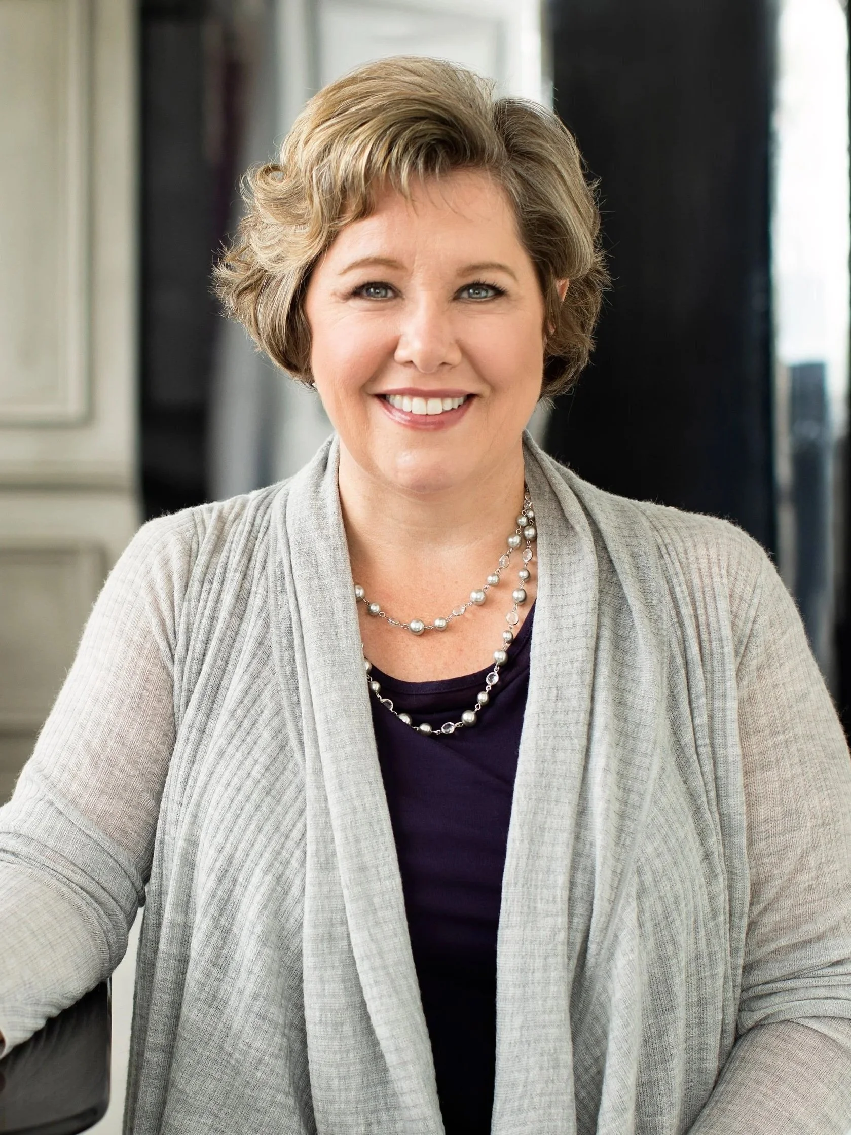 A woman with short, wavy blonde hair smiling at the camera, wearing a gray cardigan, a purple top, and a pearl necklace, seated in a room with windows.