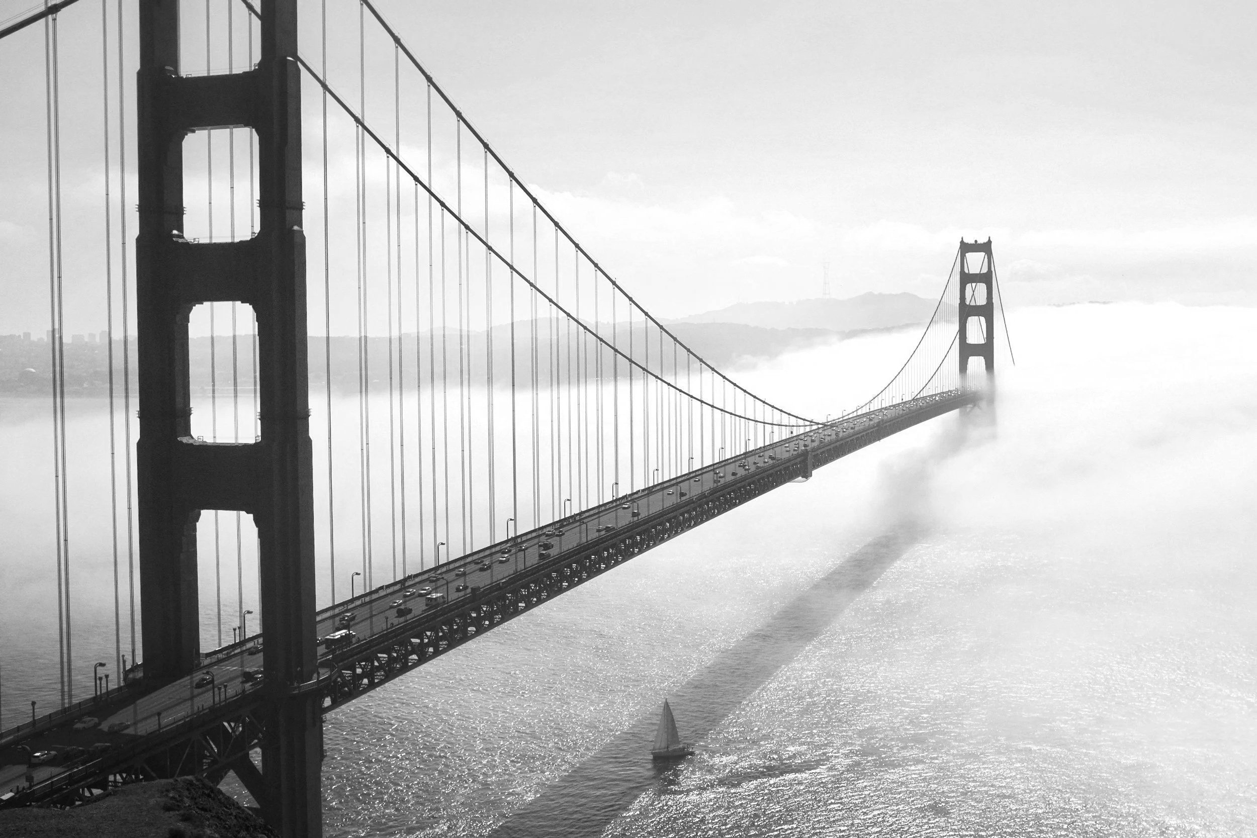 Black and white photo of the Golden Gate Bridge extending over the water with a sailboat sailing underneath.
