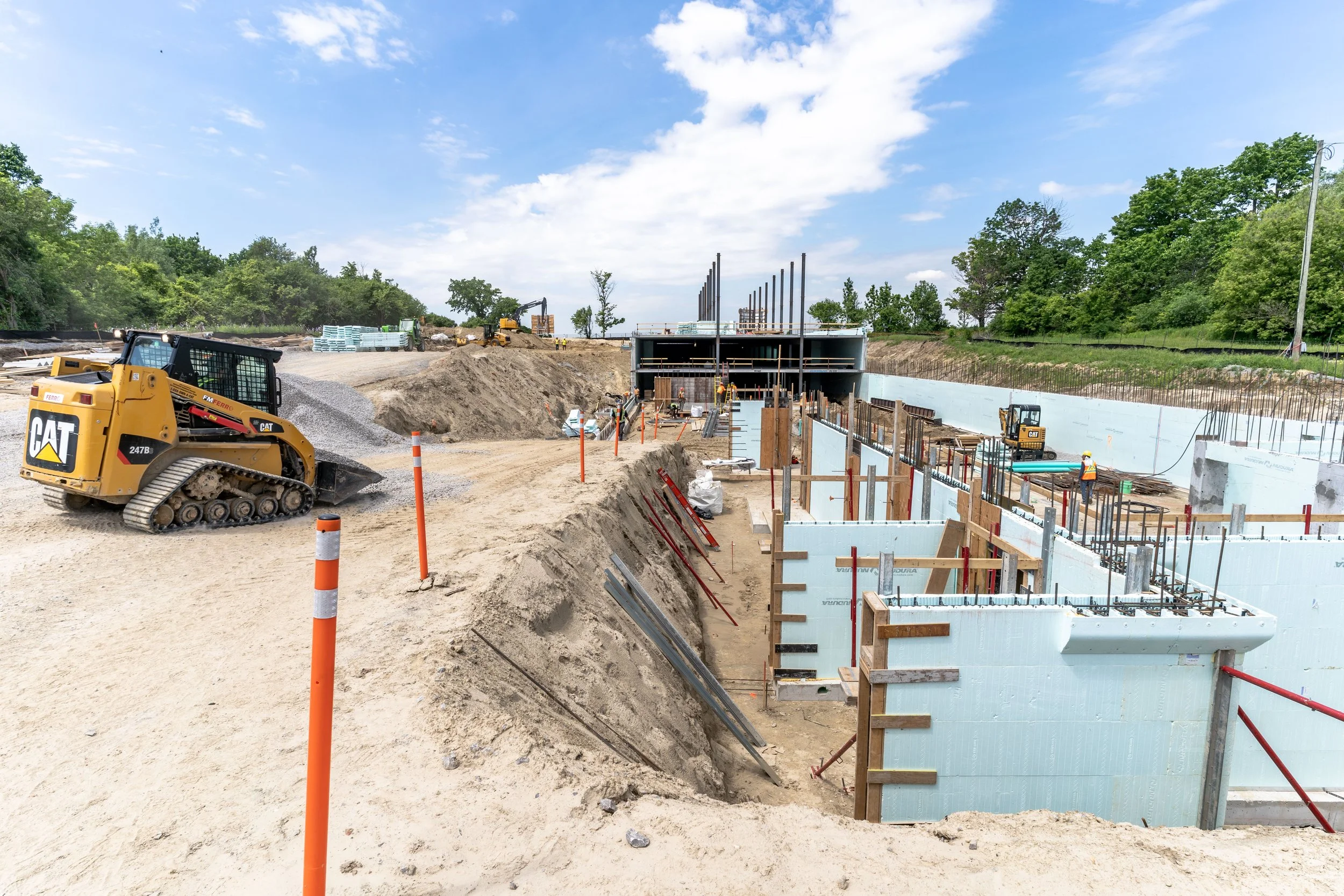 Construction site with ICF product, heavy machinery, workers, and a partially built structure with ICF concrete forms, surrounded by trees and blue sky.