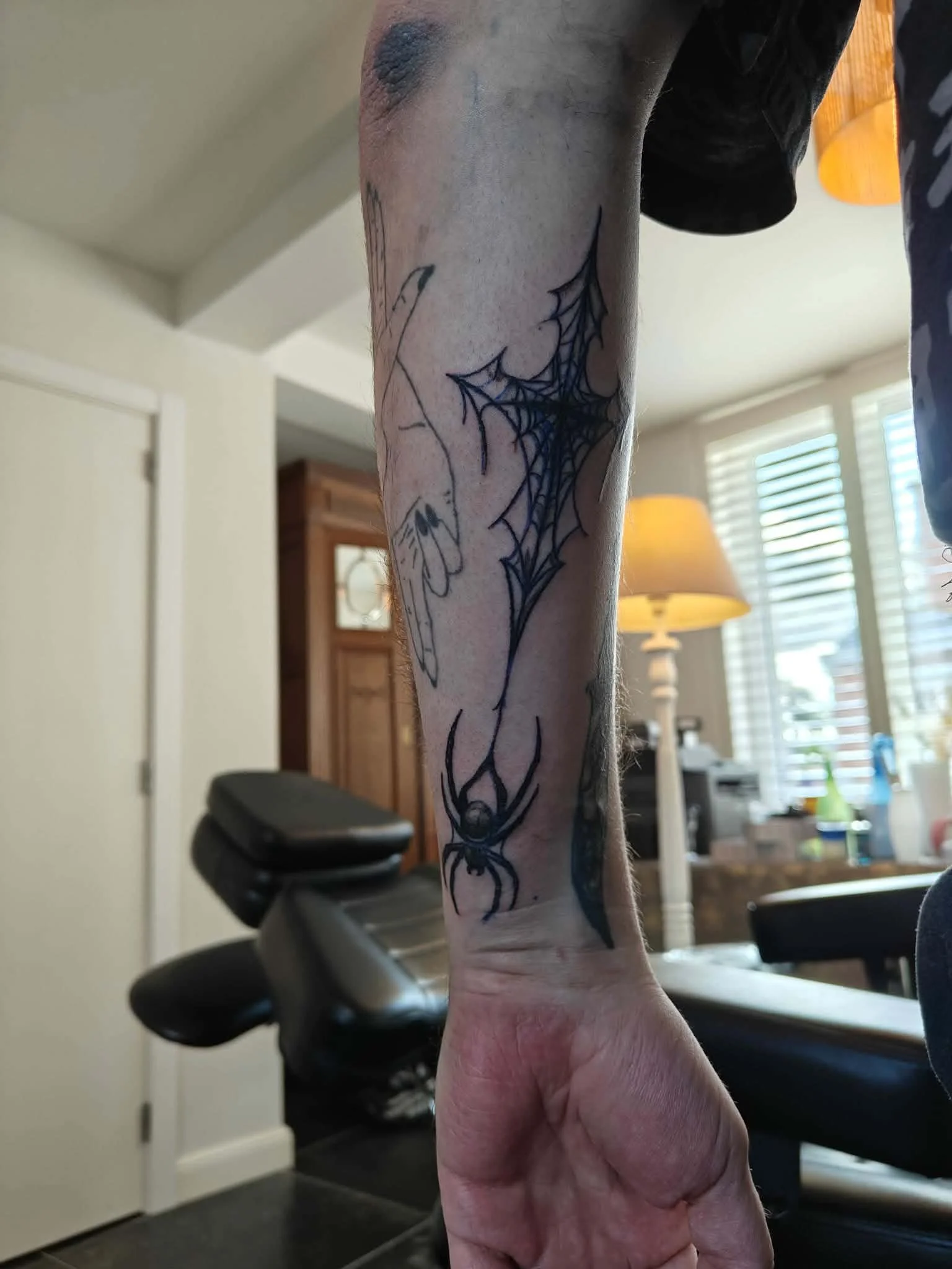 A person's arm with a tattoo of a spider and a web, holding a remote control in a room with blinds, lamps, and furniture in the background.