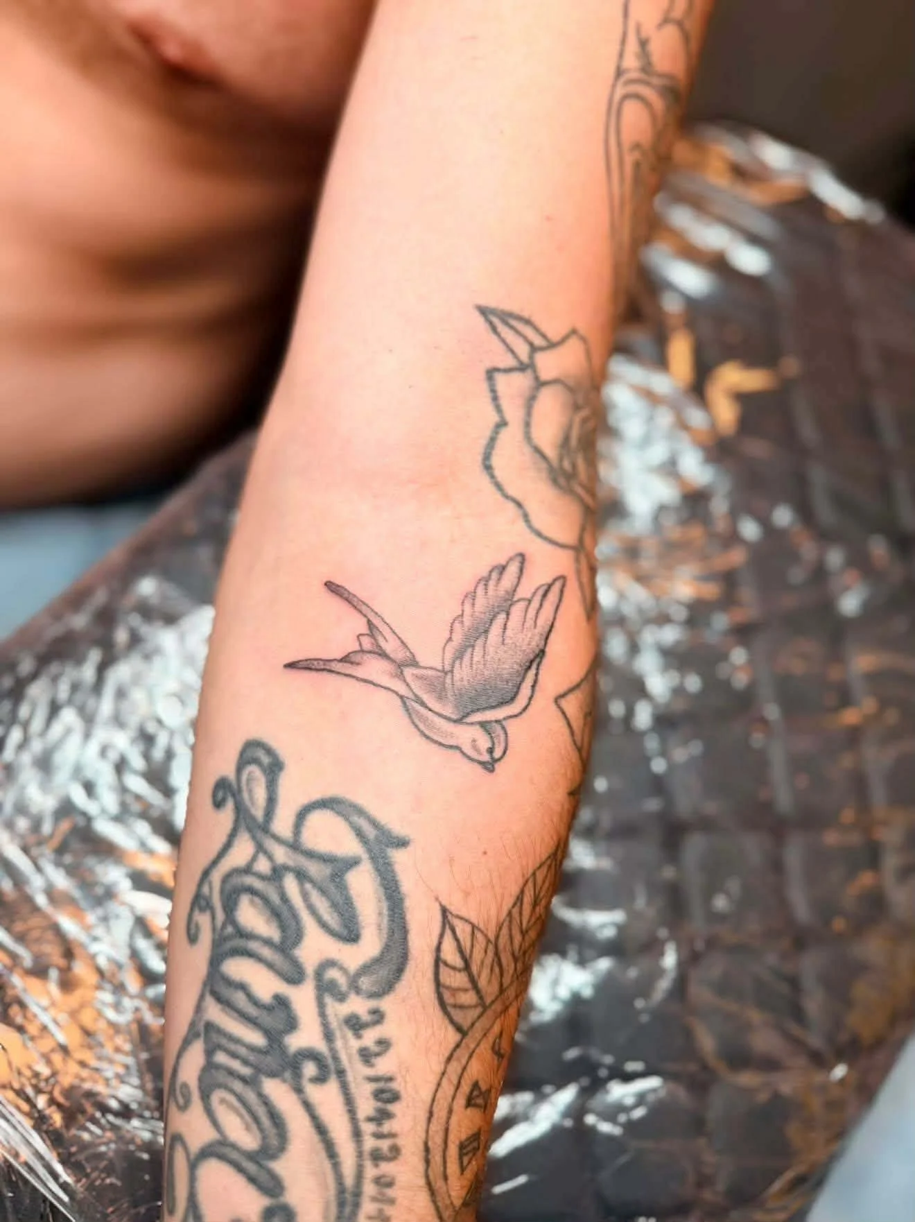 Close-up of a person's arm with black ink tattoo designs, including a rose flower and leaves. The person is shirtless, and the background shows a black surface with silver speckles.