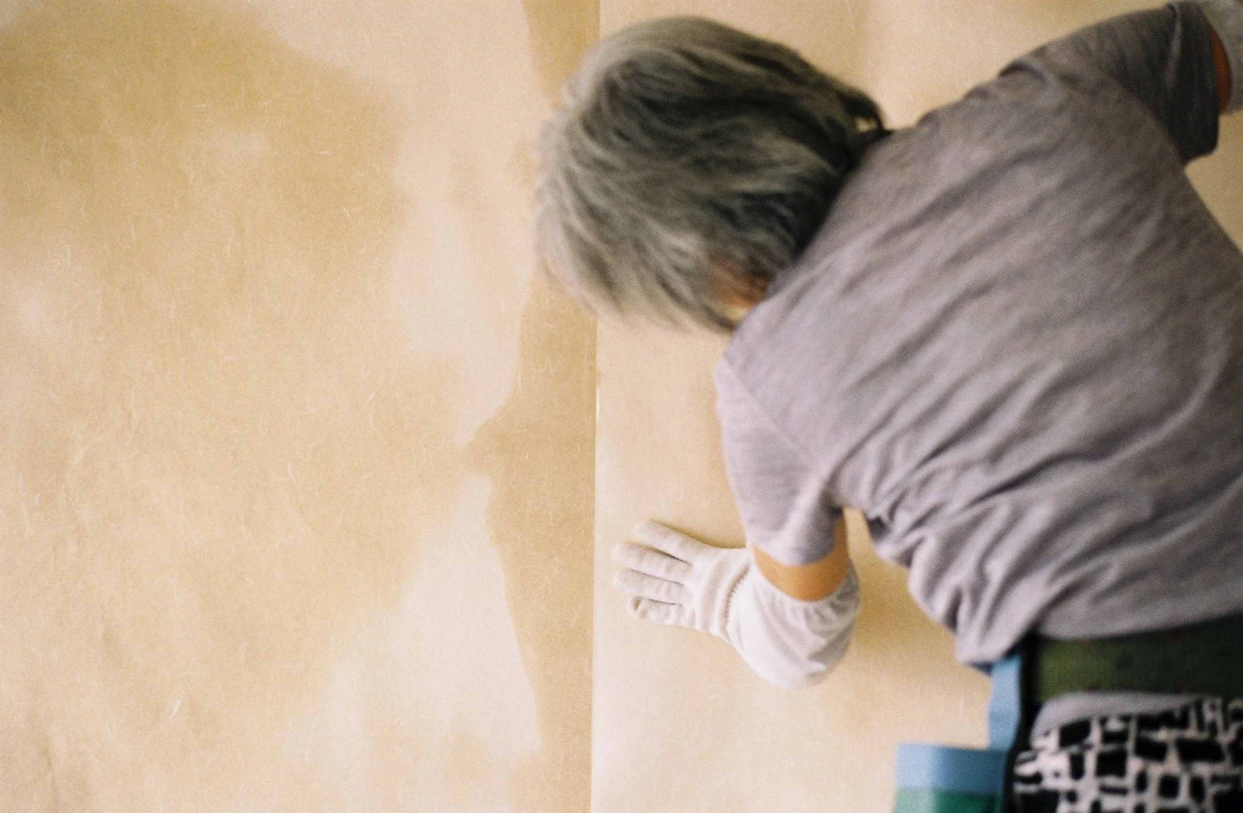 Person lying on the ground with head down, wearing a gray long-sleeved shirt and gloves, against a beige wall.