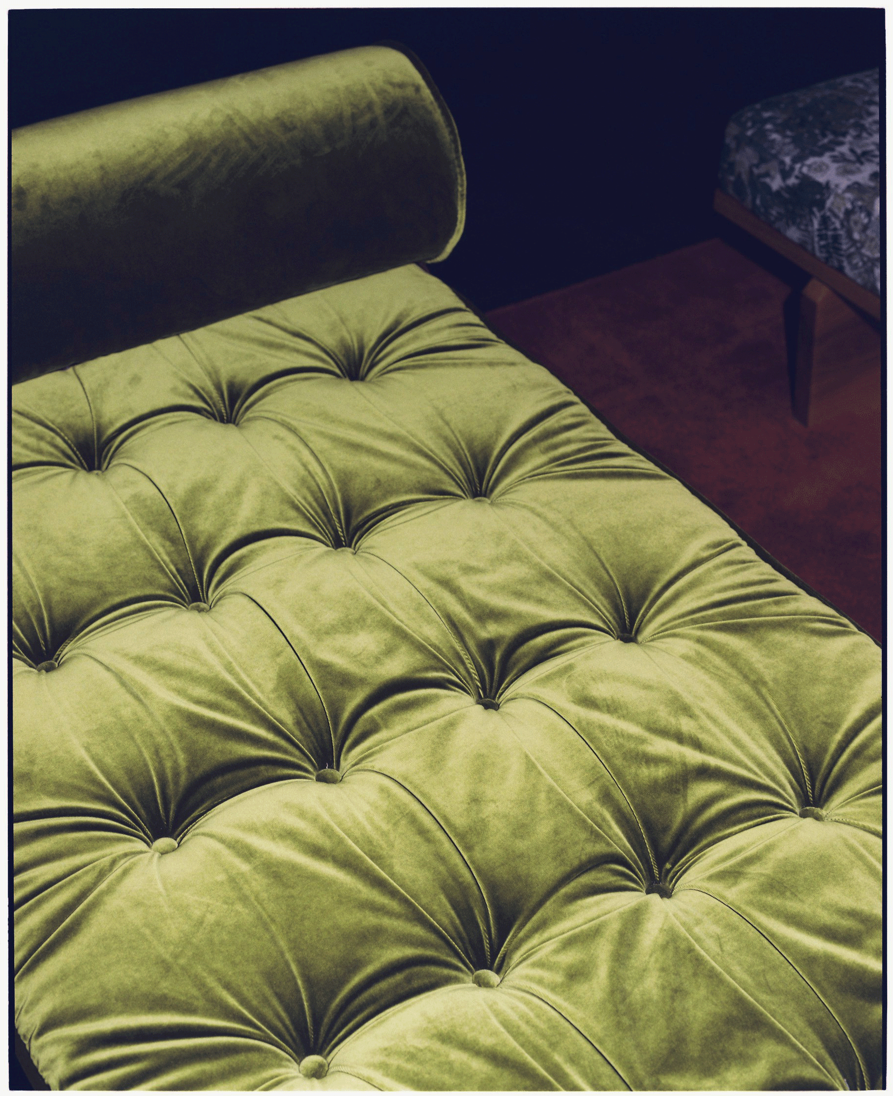 A close-up of a vintage-style, tufted, green velvet sofa with buttons, alongside a wooden side table with a cushioned top, near a dark wall.