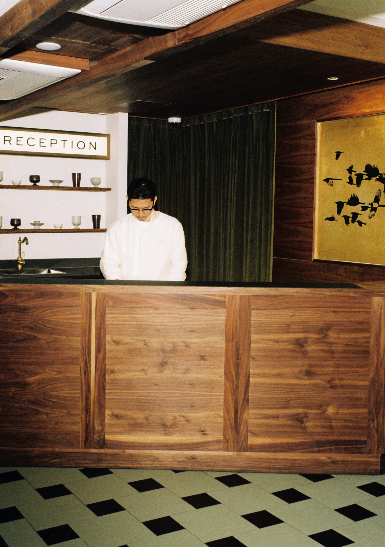 View of Seoul boutique hotel reception area with a wooden counter, a person wearing glasses behind the counter, a sign reading 'RECEPTION,' and a decorative wall art of black birds on a gold background.