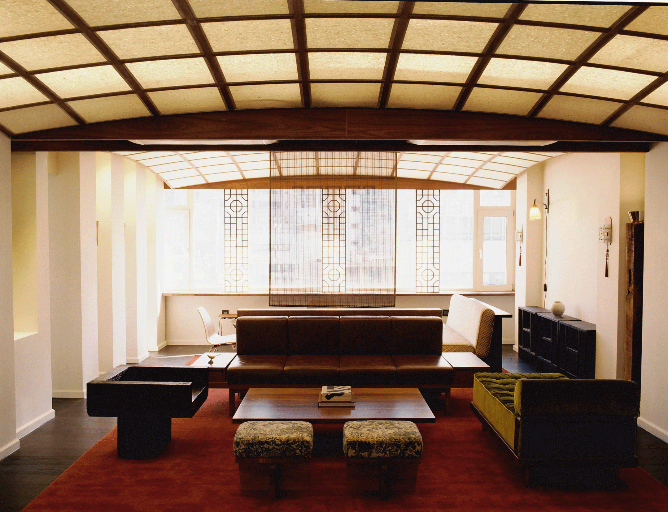 Interior of a living room with a high, grid-patterned ceiling, a large window with decorative grid details, and various furniture including a leather sofa, a yellow armchair, coffee tables, and ottomans.