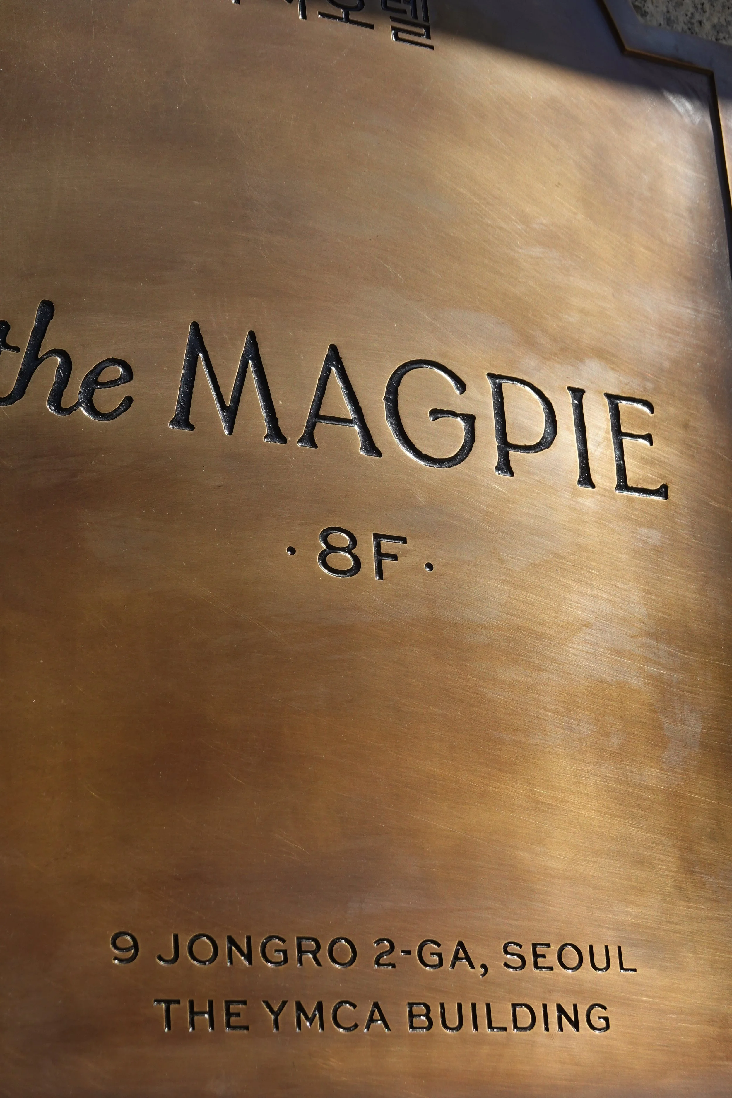 Close-up view of a bronze plaque with engraved text, including the words 'the MAGPIE' and an address, located on the YMCA building in Seoul.