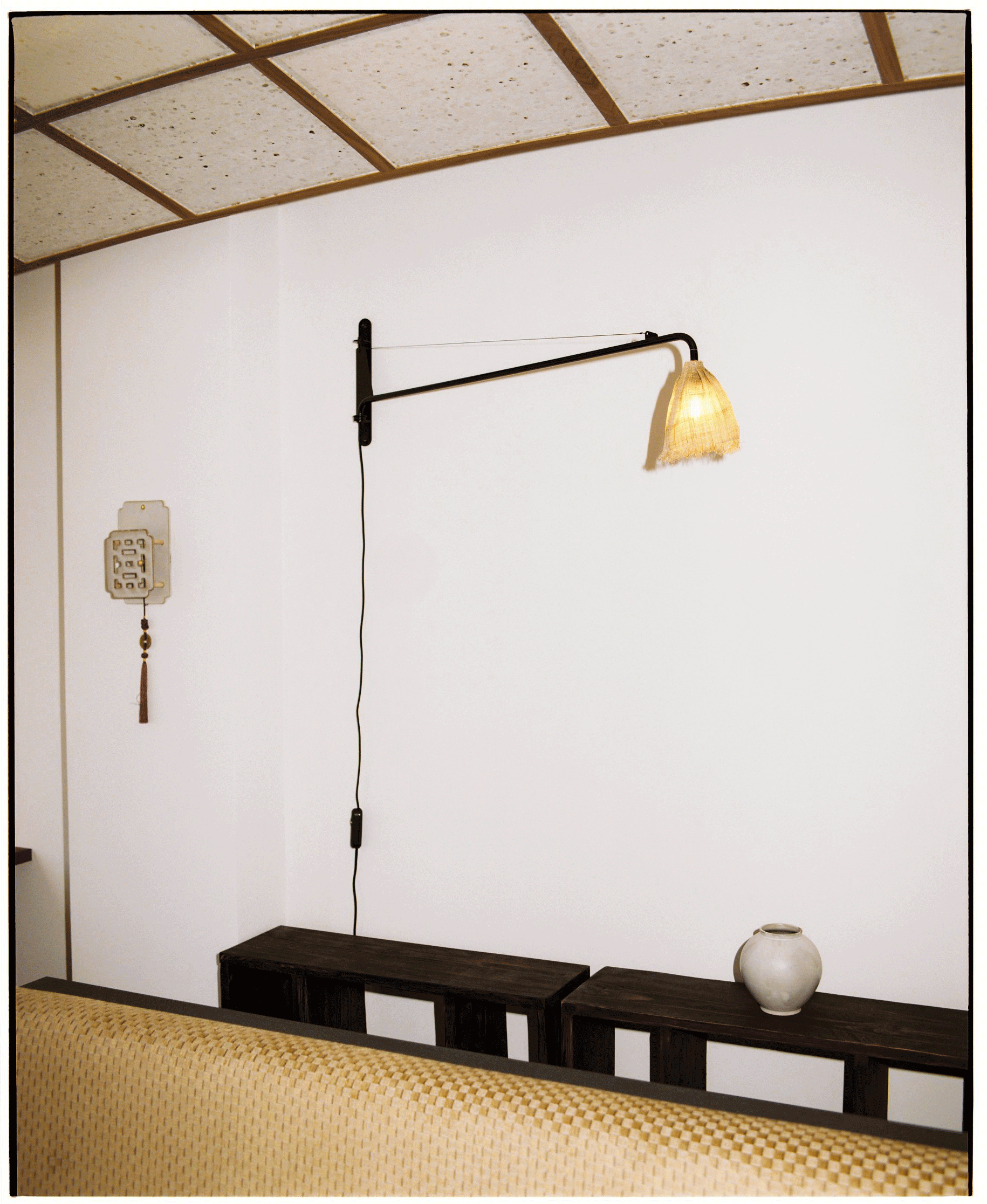 Interior room featuring a white wall with a hanging lamp, a wooden cabinet, a decorative vase, and woven textured furniture with a patterned cushion.