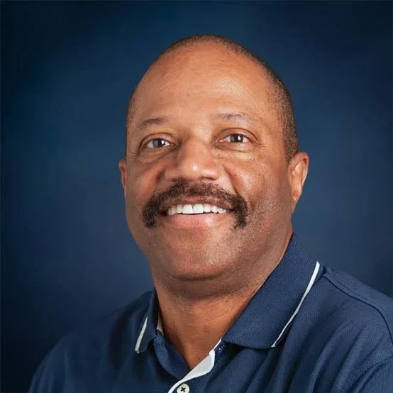 Portrait of a smiling middle-aged man with a mustache, wearing a blue polo shirt, against a dark background.