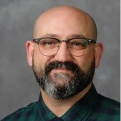Headshot of a man with glasses, a beard, and a mustache, wearing a dark plaid shirt against a gray background.