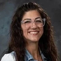 A woman with long curly hair wearing glasses and a collared shirt, smiling at the camera.