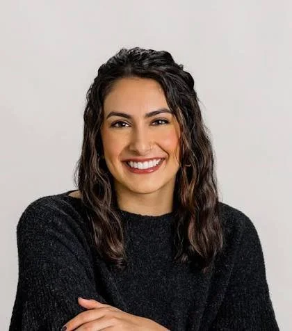 Portrait of a smiling woman with curly dark hair wearing a black sweater, standing against a plain light gray background.