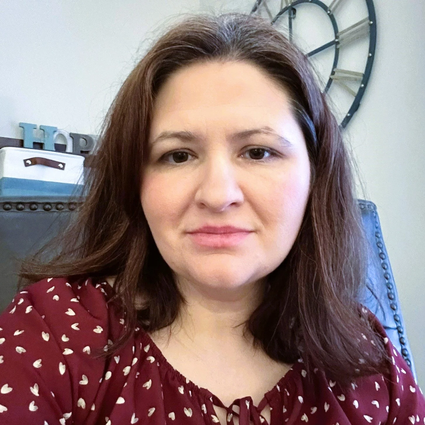 A woman with brown hair and a maroon blouse with white hearts, sitting on a leather chair, with a decorative wall clock in the background.