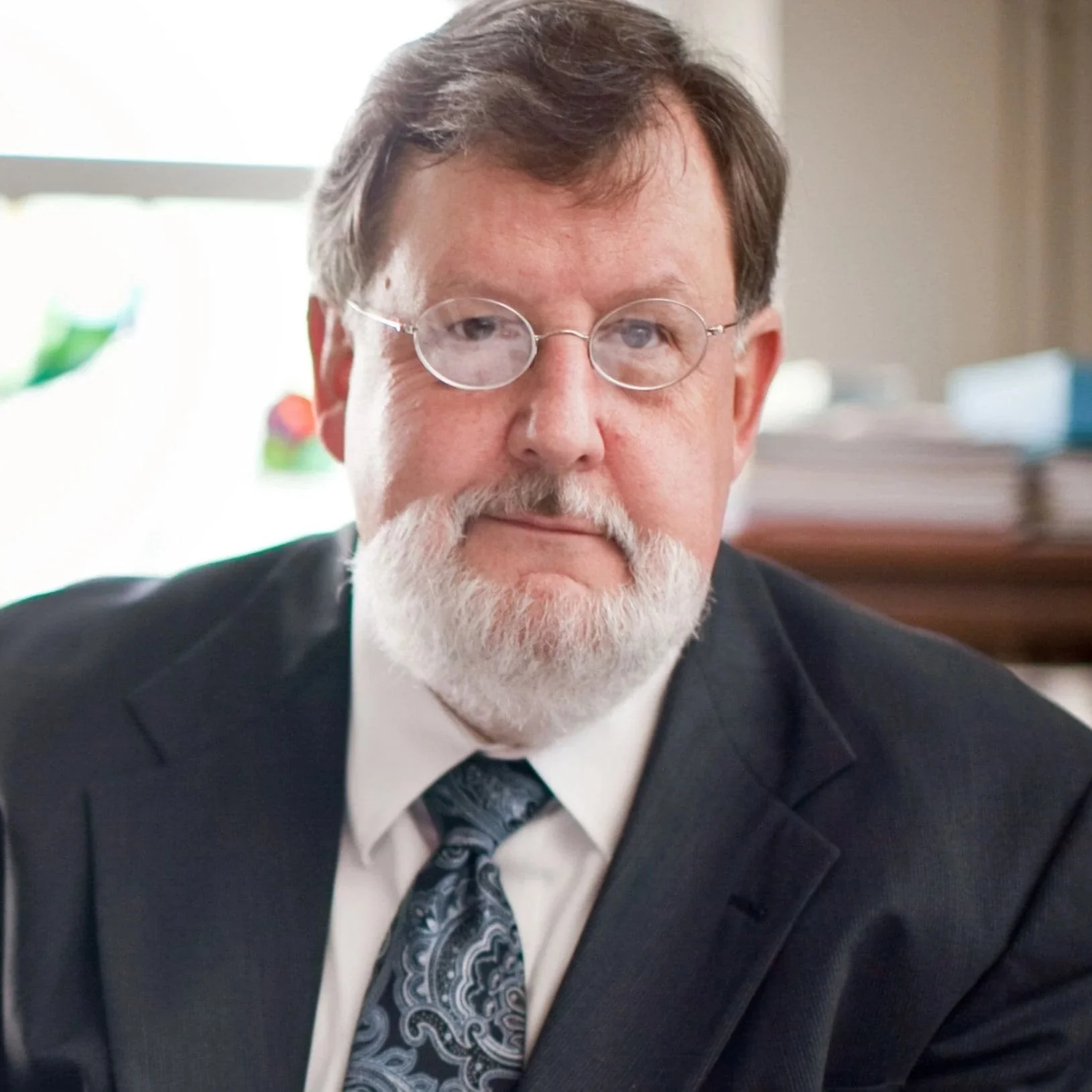 A man in a business suit with glasses, a white beard, and a patterned tie, sitting indoors during daytime.