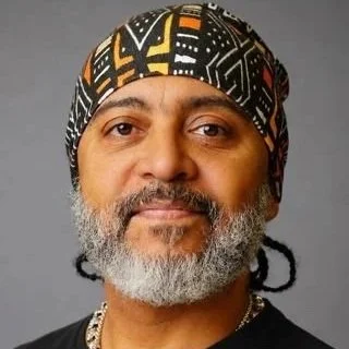 Close-up of a man with a beard wearing a patterned headscarf and a chain, against a gray background.