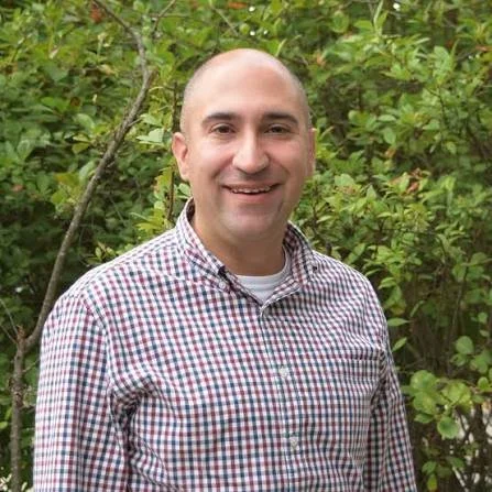 A man with a shaved head and wearing a checkered shirt standing outdoors in front of green foliage.
