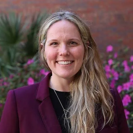 A smiling woman with long blonde hair wearing a dark blazer and a necklace, standing outdoors with pink flowers and a brick wall in the background.