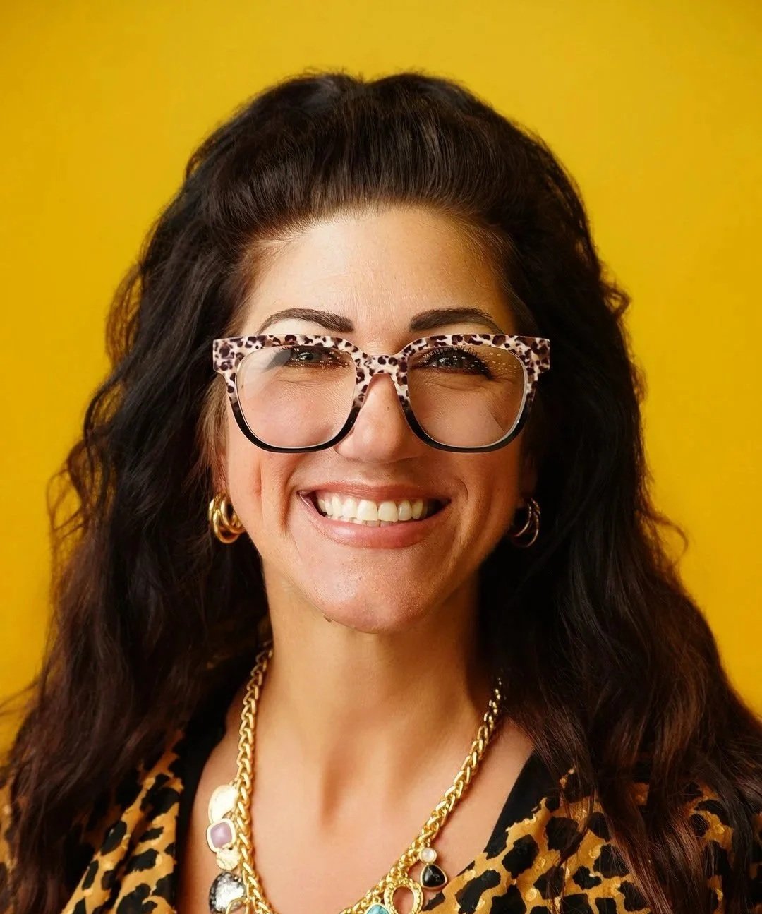 A woman with long curly hair wearing glasses and a collared shirt, smiling at the camera.
