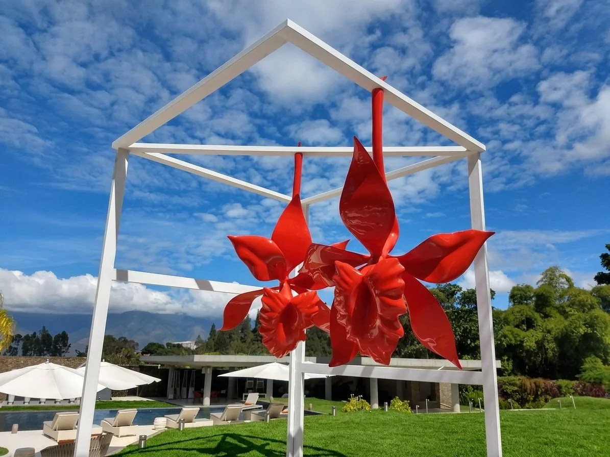 Red metal sculpture of abstract flowers under a white geometric frame, outdoor pool area with lounge chairs and umbrellas, surrounded by greenery and mountains in the background.