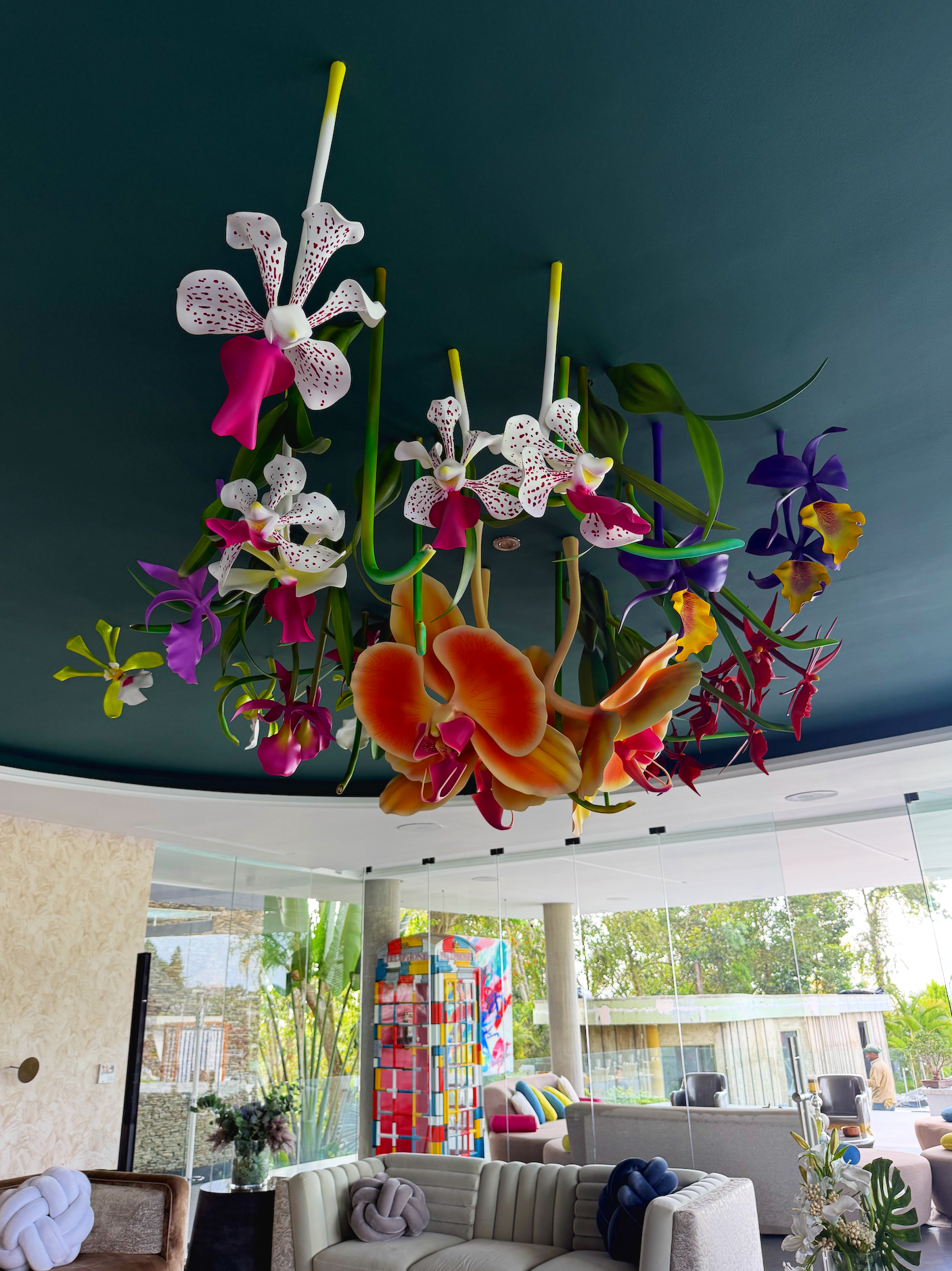 A large, colorful floral chandelier with various orchids and flowers hanging from a dark ceiling in a modern, well-lit lounge area with sofas and glass walls.