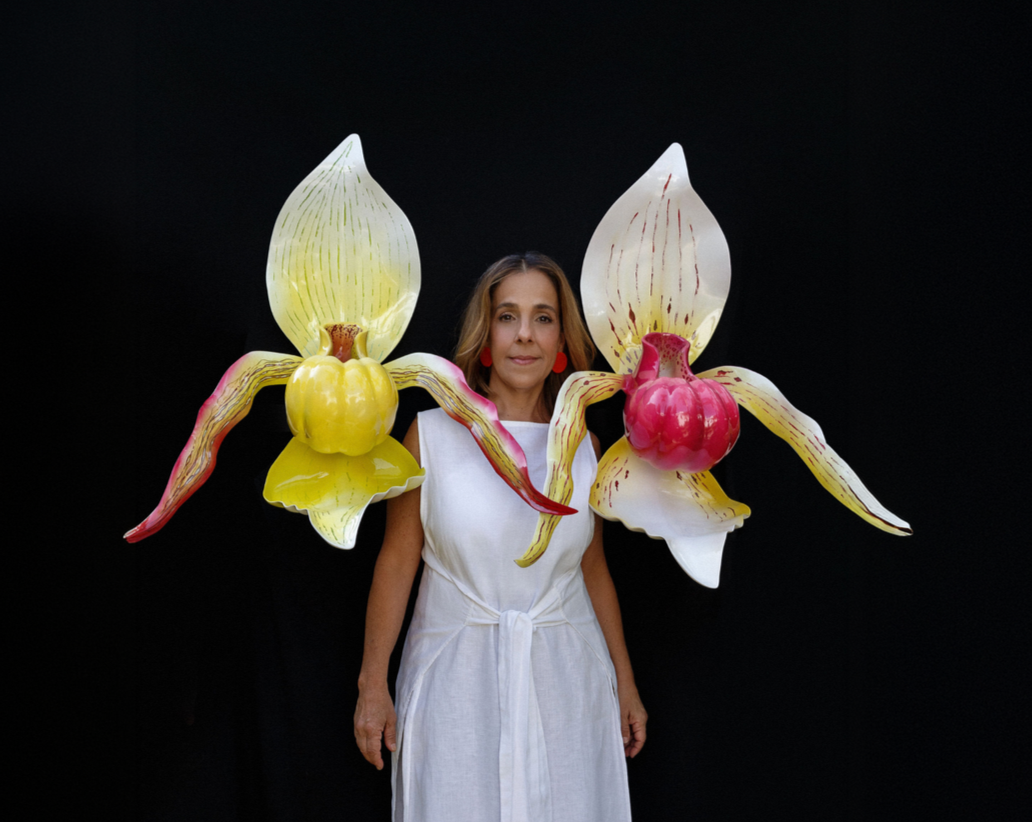 Woman in a white dress standing in front of a black background with large, colorful orchid flower sculptures around her shoulders.