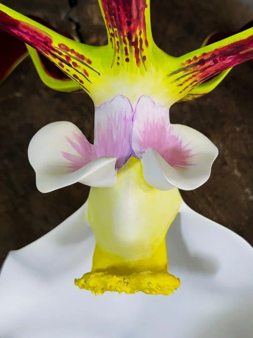 A floral sculpture resembling an animal with colorful petals and leaves, featuring yellow, pink, white, and purple hues, set on a white base against a dark background.
