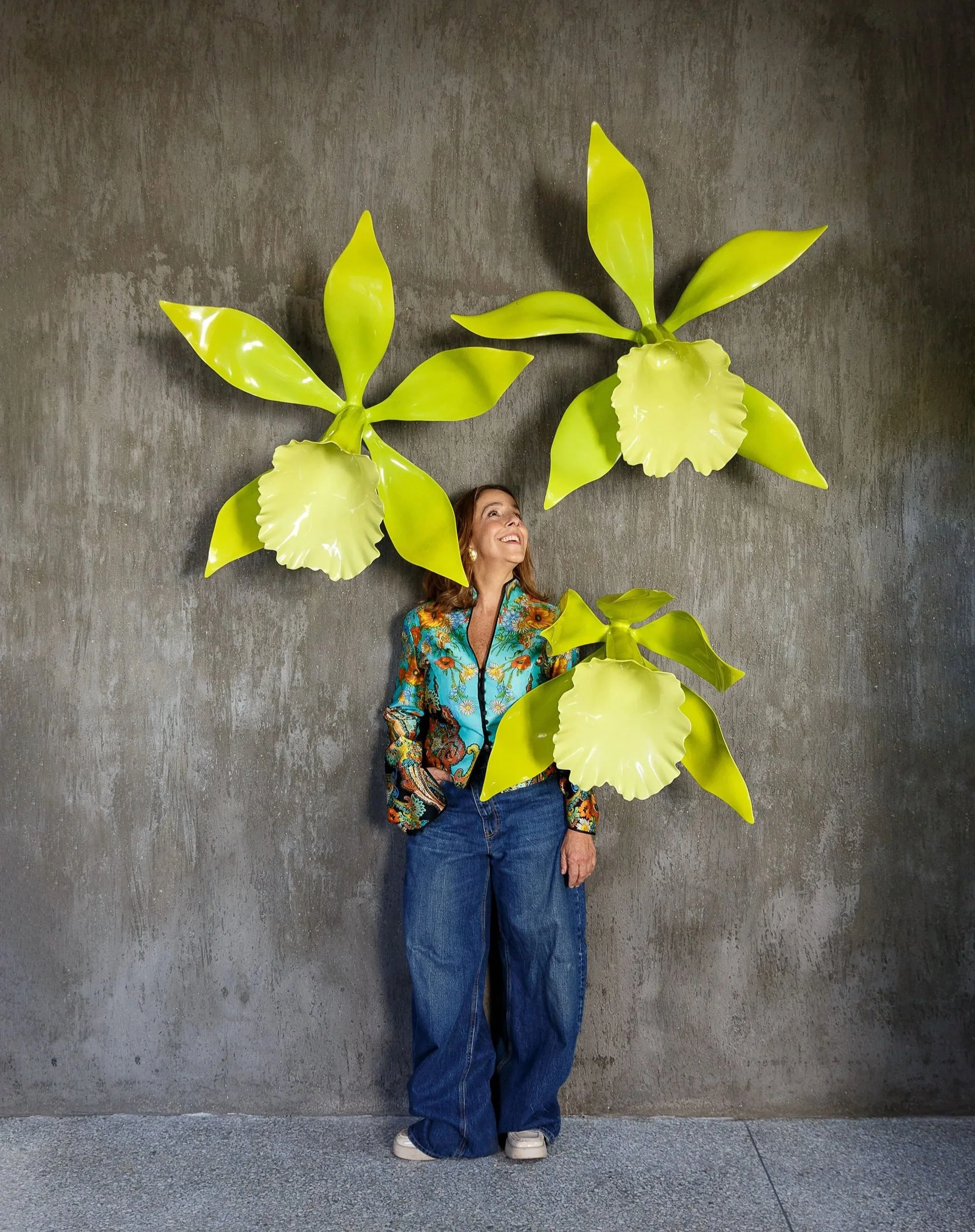 A woman with curly hair, wearing a colorful patterned jacket and blue jeans, standing against a gray wall decorated with large green tropical plant sculptures. She is looking up and smiling.