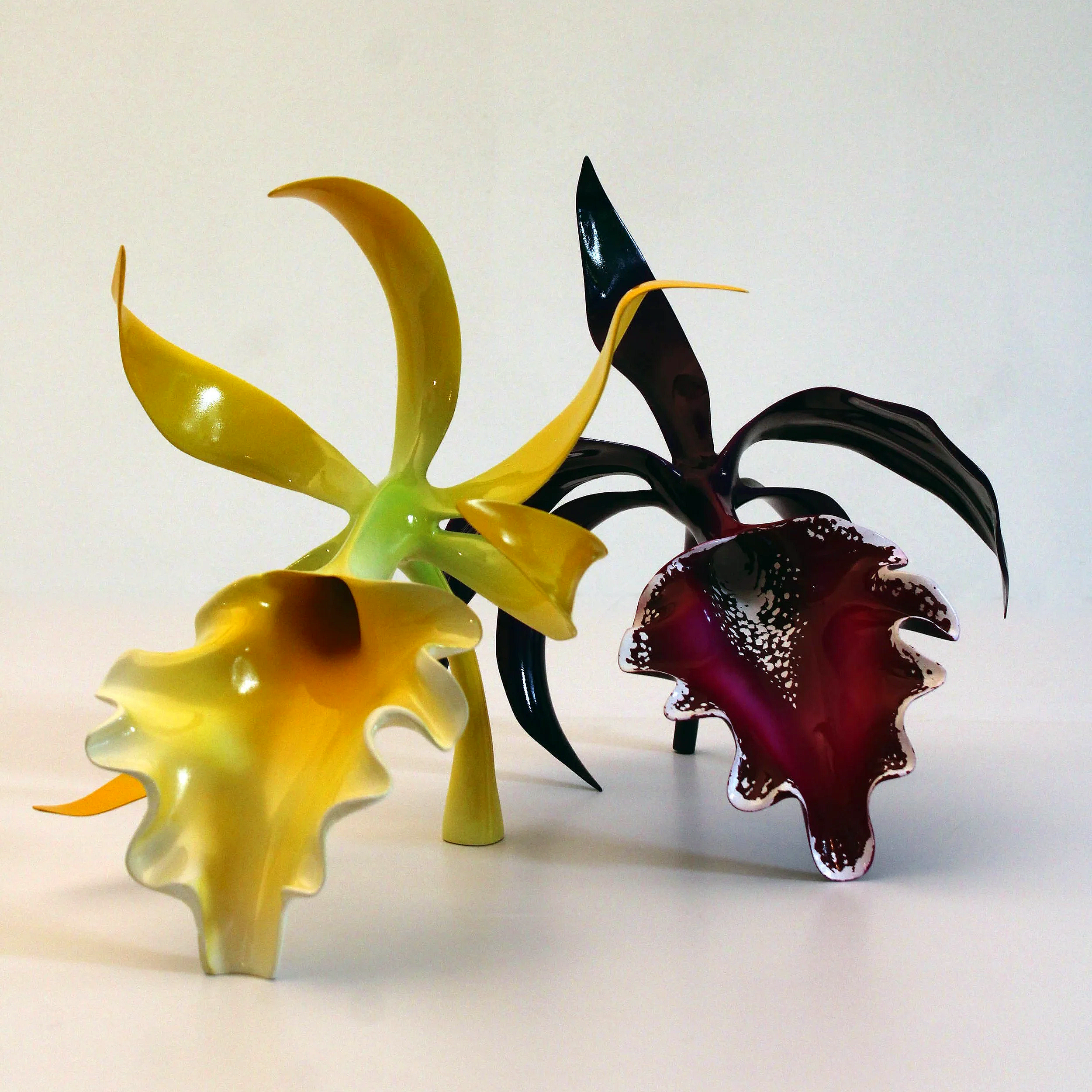 Four colorful glass sculptures resembling abstract flowers with curved, ruffled petals in yellow, black, and deep red, arranged against a plain white background.