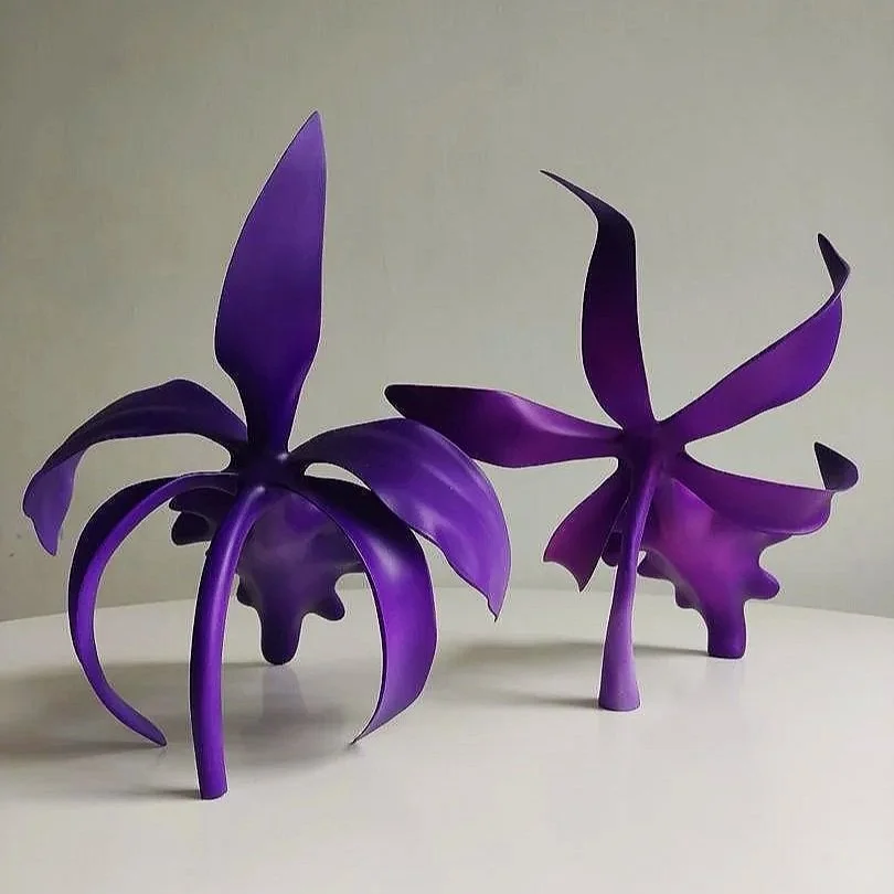 Two purple abstract sculptures resembling flowers or plants on a white surface against a plain background.