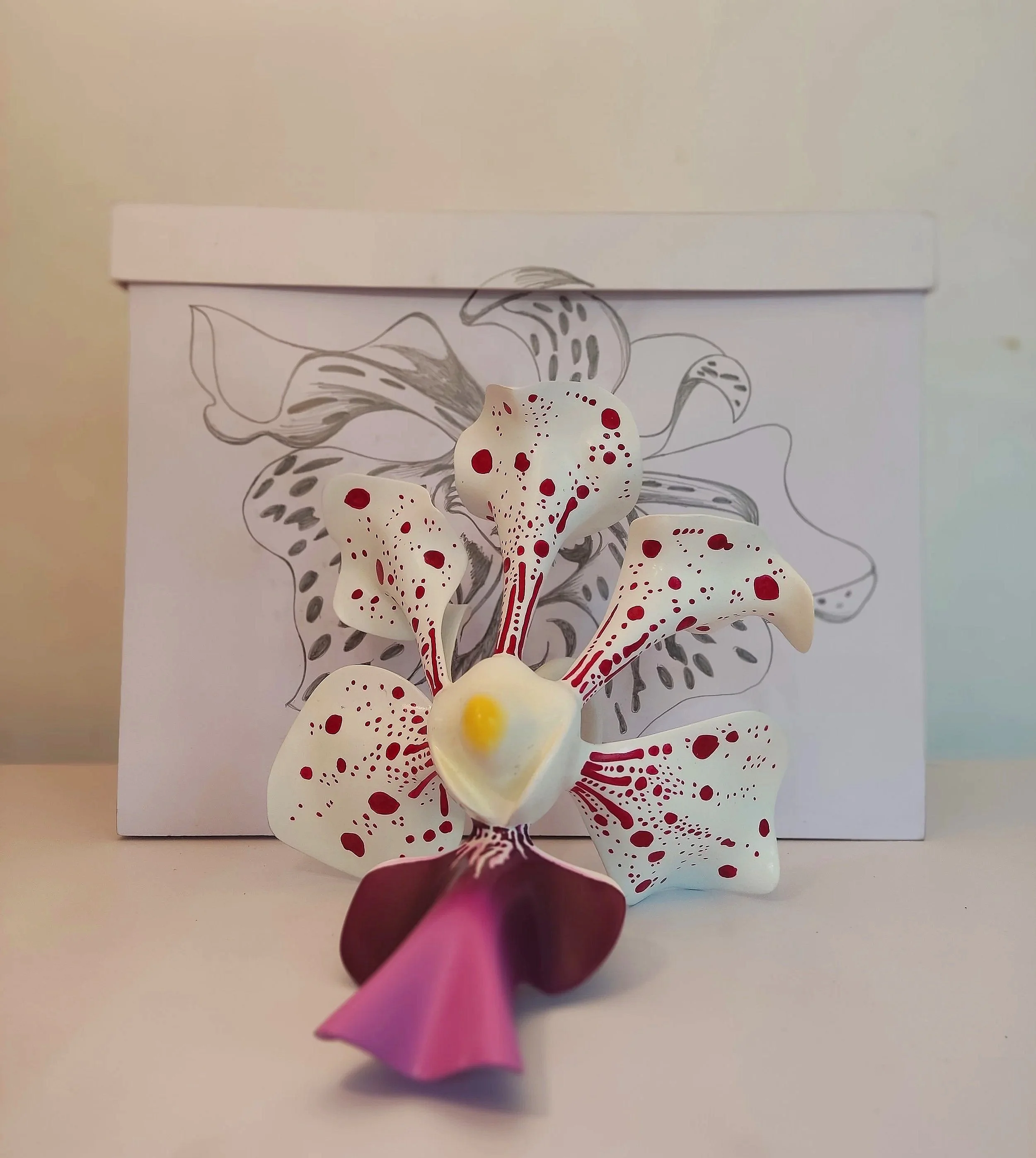 An artificial flower with pink buds and white petals with red spots in front of a black-and-white drawing of flowers and leaves.
